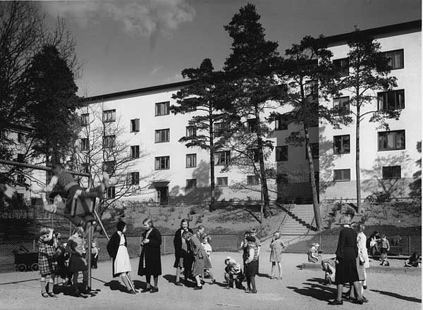 Svartvitt fotografi föreställande barn och tonåringar på lekplats framför ett flerfamiljshus